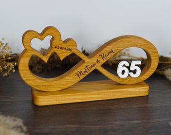 May include: Wooden infinity symbol with a heart-shaped design and the number 65. The symbol is engraved with the names "Martina & Reiner" and the date "01.05.1996". The wooden piece is set on a rectangular base.