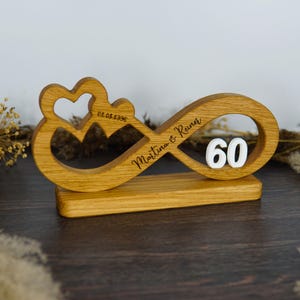 Op de afbeelding: Houten oneindigheidssymbool met een hart en het getal 60. Het symbool is gegraveerd met de namen "Martina & Reiner" en de datum "01.08.1996". De basis is een rechthoekig houten blok.