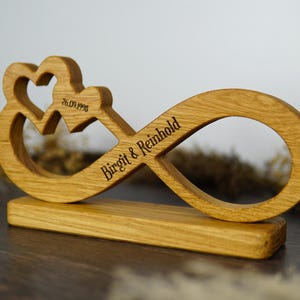 Puede incluir: Un símbolo de infinito de madera con dos corazones y una base. La fecha "26.09.1998" está grabada sobre los corazones, y los nombres "Birgit & Reinhold" están grabados en el símbolo del infinito. La madera es de color marrón claro natural.