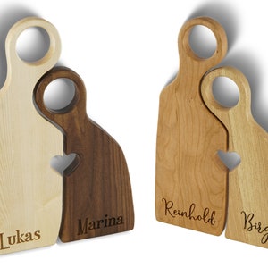 May include: Two wooden cutting boards shaped like people, with a heart cutout where they connect. The boards are personalized with the names "Lukas", "Marina", "Reinhold", and "Birgit".