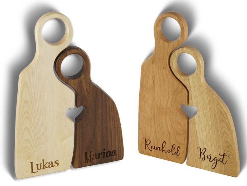May include: Two wooden cutting boards shaped like people, with a heart cutout where they connect. The boards are personalized with the names "Lukas", "Marina", "Reinhold", and "Birgit".