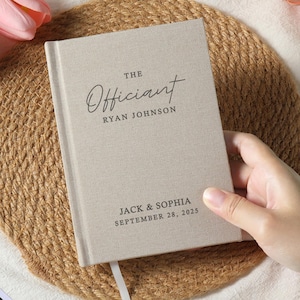 Peut inclure: Un livre beige recouvert de lin avec les mots "The Officiant" et "Ryan Johnson" imprimés sur la couverture. Le livre comprend également les noms "Jack & Sophia" et la date "September 28, 2025". Un signet en ruban blanc est visible.