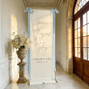 Pode incluir: Uma faixa de casamento branca com as palavras "Forever Begins Today" em escrita elegante, acentuada com fitas azul claro. A faixa inclui os nomes "Jone &hearts; Sarah" e a data "June 13, 26". Um arranjo floral em uma urna decorativa est&aacute; &agrave; esquerda.