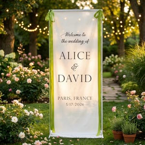 Pode incluir: Uma placa de boas-vindas de casamento branca com fitas verdes, exibindo os nomes "ALICE & DAVID" e detalhes do evento. A placa est&aacute; em um jardim com flores e luzes de corda.