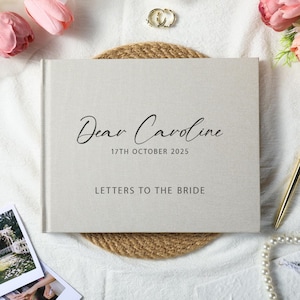Puede incluir: Un libro de visitas de boda gris claro con el texto "Dear Caroline" y "17TH OCTOBER 2025" en escritura negra. Las palabras "LETTERS TO THE BRIDE" están impresas debajo. El libro está sobre una estera tejida, con flores y un bolígrafo cerca.