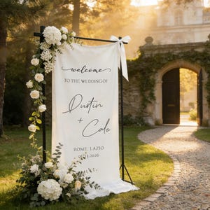 Pode incluir: Uma placa de boas-vindas de casamento branca com o texto "welcome to the wedding of Dustin + Cole" e a data 06.13.2026. A placa &eacute; decorada com flores brancas e vegeta&ccedil;&atilde;o e est&aacute; montada ao ar livre.