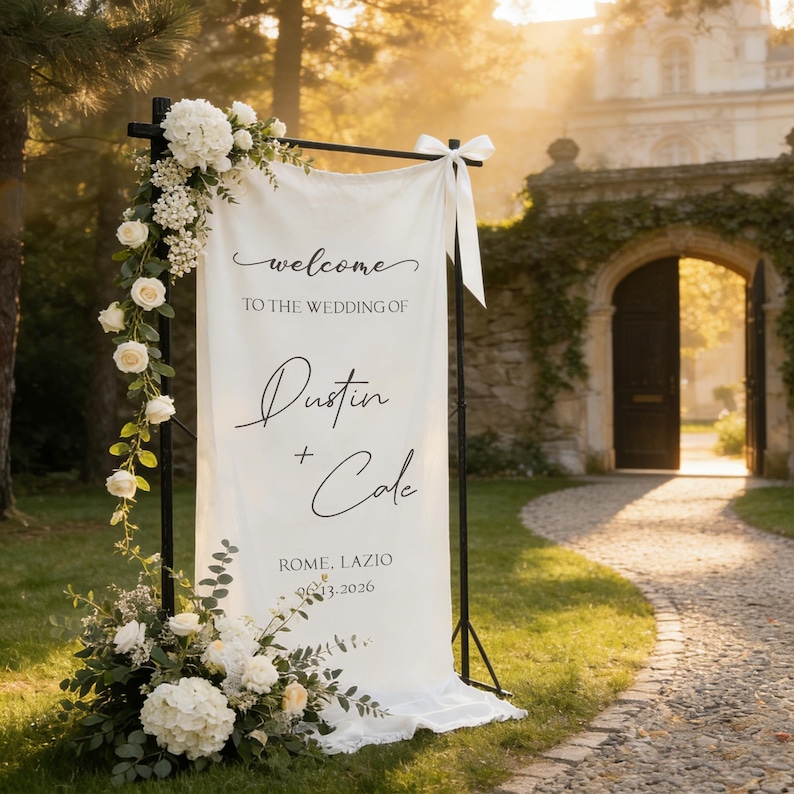 Pode incluir: Uma placa de boas-vindas de casamento branca com o texto "welcome to the wedding of Dustin + Cole" e a data 06.13.2026. A placa &eacute; decorada com flores brancas e vegeta&ccedil;&atilde;o e est&aacute; montada ao ar livre.