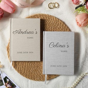 Puede incluir: Dos libros de votos con tapa dura de tela. El libro de la izquierda es beige con el texto negro Andrea's VOWS JUNE 26TH 2026. El libro de la derecha es gris claro con el texto negro Celina's VOWS JUNE 26TH 2026. Ambos libros incluyen un marcapáginas.