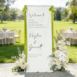Pode incluir: Uma placa de boas-vindas de casamento branca com fitas verdes e arranjos florais. A placa diz "Welcome to Our Happily Ever After" e apresenta os nomes Kaylee & Garrett, juntamente com o local e a data.