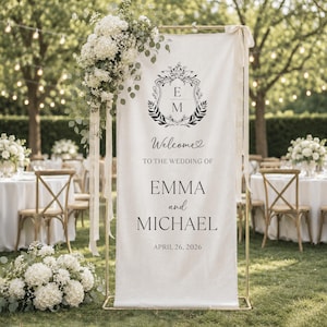 May include: A tall, rectangular off-white fabric wedding welcome sign suspended on a gold metal frame. The sign features a floral crest with the initials E M, the text Welcome to the wedding of Emma and Michael, and the date April 26, 2026.