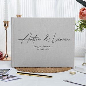 May include: A light grey linen wedding guest book with the names "Austin & Lauren" in elegant script. The location "Prague, Bohemia" and the date "14 may 2026" are printed below the names. The book is on a woven mat.