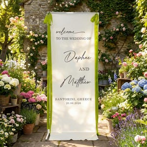 Pode incluir: Uma placa de boas-vindas de casamento branca com fitas verdes, com os nomes de Daphne e Matthew, e a localiza&ccedil;&atilde;o Santorini, Gr&eacute;cia, com a data 20.06.2026. A placa est&aacute; rodeada por flores e vegeta&ccedil;&atilde;o exuberantes.