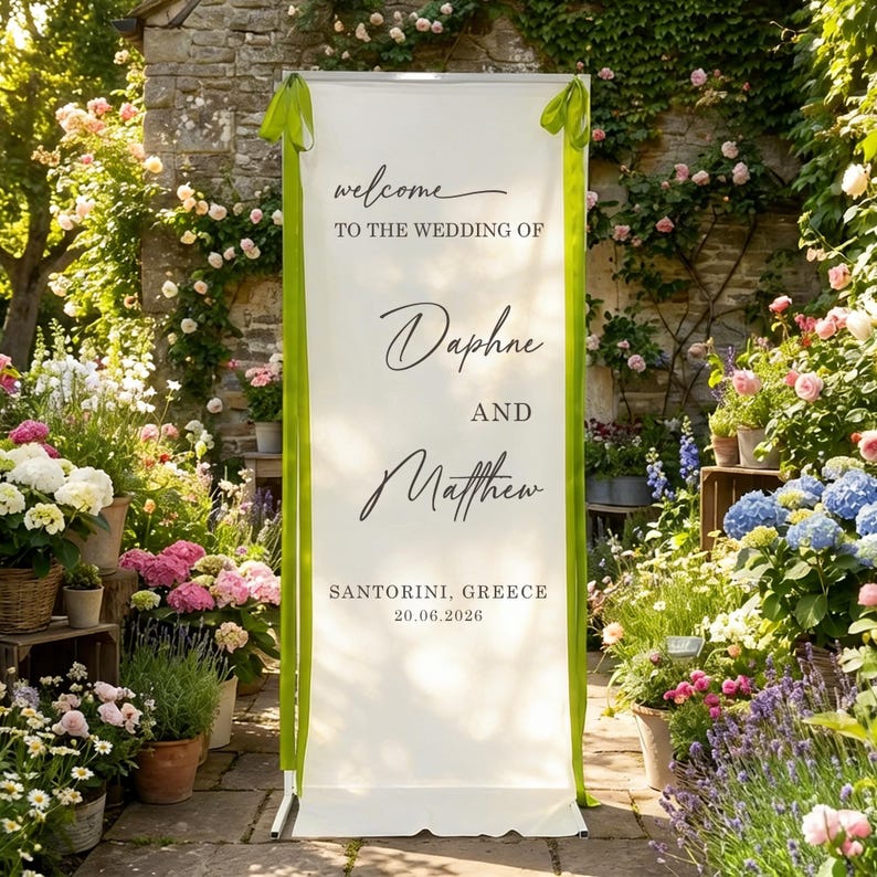 Pode incluir: Uma placa de boas-vindas de casamento branca com fitas verdes, com os nomes de Daphne e Matthew, e a localiza&ccedil;&atilde;o Santorini, Gr&eacute;cia, com a data 20.06.2026. A placa est&aacute; rodeada por flores e vegeta&ccedil;&atilde;o exuberantes.