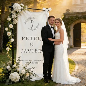 Pode incluir: Um casal posa em frente a uma placa de casamento. A placa diz "Peter & Javier" com a data "4 de junho de 2026" e a localiza&ccedil;&atilde;o "Barcelona, Catalunha". A placa &eacute; decorada com arranjos florais e vegeta&ccedil;&atilde;o. O casal est&aacute; vestido com trajes formais de casamento.