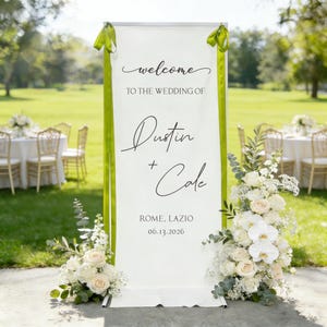 Pode incluir: Uma placa de boas-vindas de casamento branca com fitas verdes e arranjos florais. A placa diz "welcome to the wedding of Dustin + Cole" com a localiza&ccedil;&atilde;o e a data abaixo. A placa &eacute; cercada por flores brancas e verdes.