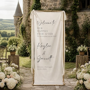 May include: A tall, off-white fabric banner suspended from a gold metal frame. The banner features black printed text that reads: Welcome to OUR HAPPILY EVER AFTER We are so glad you are here! Kaylee & Garrett Óbidos, Portugal 16.05.2026.