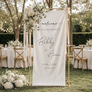 Peut inclure: Une pancarte de bienvenue de mariage en tissu blanc suspendue sur un cadre en métal doré. Le texte indique : welcome TO THE WEDDING OF Bobby + Anna ROME, LAZIO 13.06.2026. La pancarte est ornée d'un grand arrangement d'hortensias blancs et d'eucalyptus.