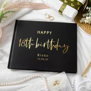 Op de afbeelding: Een zwart hardcover boek met gouden letters met de tekst "HAPPY 18th birthday" en de naam "Rieke" met de datum "12.04.26". Het boek is omgeven door een wit oppervlak, een geschenkdoos en sieraden.