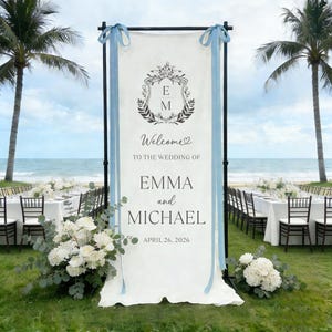 Pode incluir: Uma placa de boas-vindas de casamento branca com os nomes Emma e Michael e a data de 26 de abril de 2026. A placa &eacute; decorada com um bras&atilde;o floral e fitas azul claro. A placa est&aacute; montada numa praia com palmeiras.