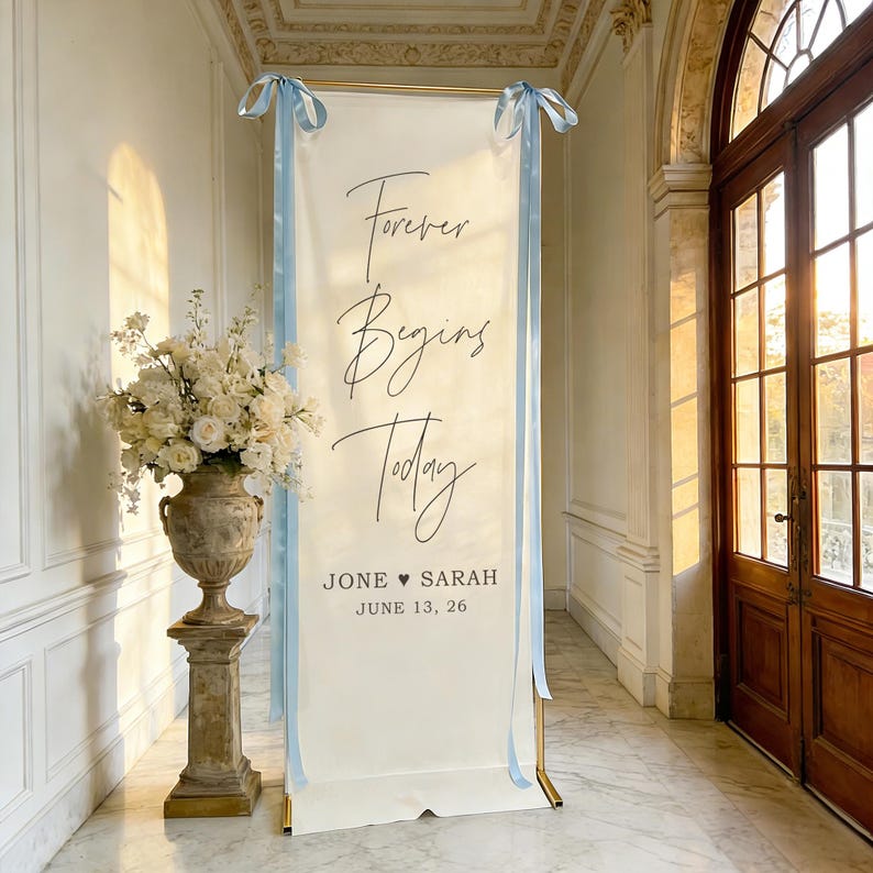 Pode incluir: Uma faixa de casamento branca com as palavras "Forever Begins Today" em escrita elegante, acentuada com fitas azul claro. A faixa inclui os nomes "Jone &hearts; Sarah" e a data "June 13, 26". Um arranjo floral em uma urna decorativa est&aacute; &agrave; esquerda.