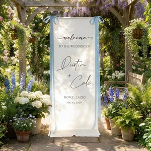 Pode incluir: Uma placa de boas-vindas branca com fitas azul claro, num jardim. A placa diz: "welcome to the wedding of Dustin + Cole, Rome, Lazio, 06.13.2026." O fundo apresenta vegeta&ccedil;&atilde;o exuberante, flores e uma p&eacute;rgola de madeira.