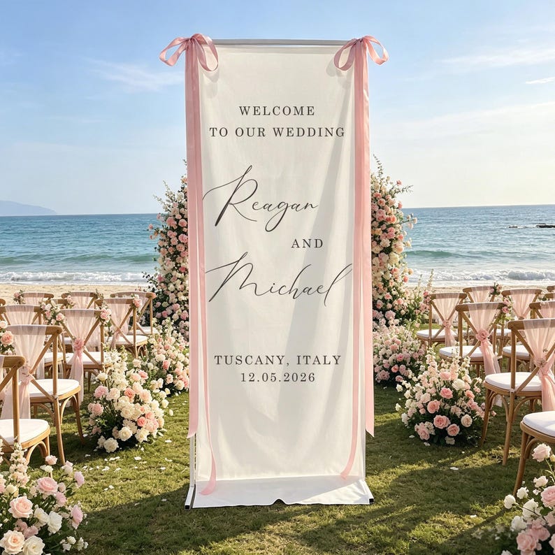 Pode incluir: Uma placa de boas-vindas de casamento branca com fitas rosa, com os nomes "Reagan e Michael" e a localiza&ccedil;&atilde;o "Toscana, It&aacute;lia", datada de "12.05.2026". A placa est&aacute; em uma praia com decora&ccedil;&otilde;es florais.