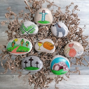 May include: Painted stones depicting scenes from the Easter story. The stones feature images of a palm branch, a donkey, a chalice and bread, a road with trees, three crosses, and a tomb.