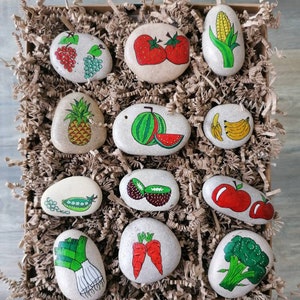 May include: A collection of painted stones featuring various fruits and vegetables, including strawberries, a pineapple, a watermelon, bananas, peas, carrots, an apple, broccoli, and a leek. The stones are arranged in a brown paper basket.