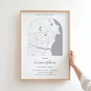 Op de afbeelding: Een ingelijste verticale wandkunstprint met een kaart in een boogvorm en een rood hartpictogram. De tekst luidt A NEW CHAPTER BEGINS, Lauren & Oliver, OUR FIRST HOME, 30 Shrigley Avenue, Ashington, 26th January 2026, 55.18483 N - 1.54099 W.
