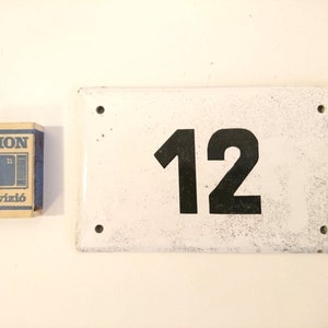 May include: White enamel house number sign with black painted number 12. The sign has two holes on each side for mounting.