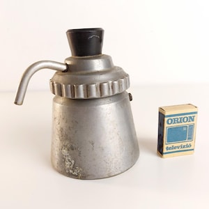 Peut inclure: Une cafetière expresso argentée vintage avec une poignée et un dessus noirs. Une petite boîte d'allumettes "ORION televizió" est à côté de la cafetière. La cafetière a une poignée et une conception nervurée au milieu.