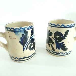 May include: Two white ceramic mugs with blue and black floral designs. The mugs have a rounded shape and a handle.