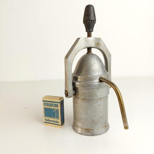 May include: A vintage aluminium stovetop coffee maker with a dome-shaped lid, a curved brass spout, and a black handle knob. Beside it sits a small rectangular matchbox with the blue text ORION and televízió printed on the front surface.