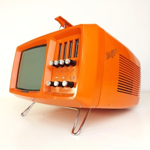 May include: A bright orange portable television set with a rectangular screen, a top handle, and two thin chrome wire legs. The front panel features four vertical sliders, four circular control knobs labelled 1, 2, 3, 4, and three additional icons.