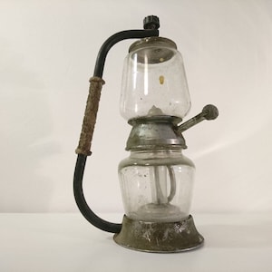 May include: A vintage glass and metal coffee maker with a black handle and a glass carafe. The coffee maker has a metal base and a glass lid.