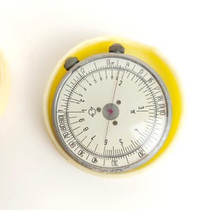 May include: A circular slide rule with a cream-colored face, black numerical markings, and a thin red indicator needle. The device is encased in a pale yellow plastic housing and features two small metallic adjustment knobs at the top edge.