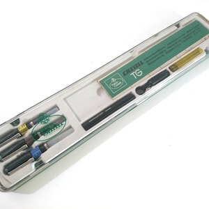 May include: A clear plastic case containing a set of Castell TG technical drawing pens. The case has a green label with the Castell logo and the text "TG". The pens are black with silver accents and have different line widths.