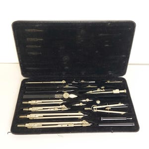 May include: A vintage drafting instrument set housed in a black rectangular case with a velvet-lined interior. The collection includes various metal compasses, dividers, and extension bars with silver-toned finishes and small adjustment knobs.