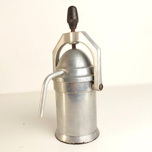 May include: A vintage, silver-toned espresso maker with a black handle. The manual coffee machine features a curved spout and a domed top, with a handle for pressing. The base is circular, and the overall design is industrial.