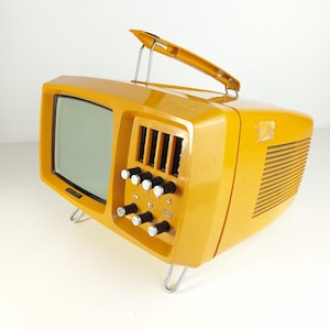 Puede incluir: Un televisor portátil de color amarillo mostaza con pantalla rectangular, asa de transporte superior y cuatro mandos de control negros con tapa plateada en el panel frontal. El dispositivo cuenta con una carcasa lateral ventilada y patas.