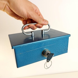 Op de afbeelding: Een blauwe metalen kassa met een zilveren handvat en een sleutel in het slot. De doos wordt door een hand vastgehouden, wat het draagbare ontwerp laat zien. De doos heeft een getextureerd oppervlak.