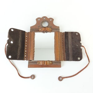 May include: A brown leather folding mirror with a decorative design. The mirror has a small, round mirror inside and a leather strap.