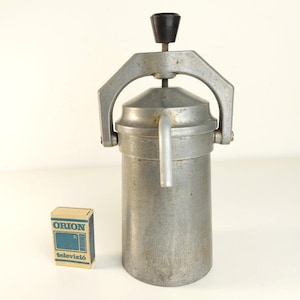 May include: A vintage silver-toned metal stovetop coffee maker with a cylindrical body, a conical lid, and a top-mounted black knob handle. Beside it sits a small rectangular matchbox with the blue text ORION and televízió printed on it.