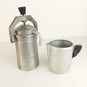 May include: A vintage aluminum stovetop espresso maker with a black knob on the top handle and a matching aluminum pitcher featuring a black handle. Both items have a metallic finish and show signs of wear consistent with regular kitchen use.