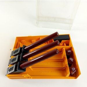 May include: An open orange plastic case containing three dark brown Rotring 800 pens. The case also holds several red pen tips with varying sizes, and a clear plastic lid is in the background. The pens are angled, and the case is on a white surface.