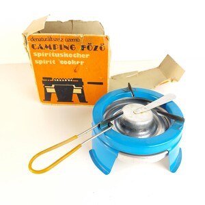 Könnte beinhalten: Ein blauer Vintage-Campingkocher mit silbernem Brenner und gelbem Griff. Der Kocher steht neben seiner originalen orangefarbenen Box mit dem Text "CAMPING FŐZŐ spirituskocher spirit cooker".