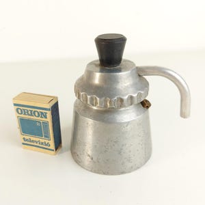 May include: A small silver-coloured metal stovetop coffee maker with a black knob on the lid and a curved handle, positioned next to a rectangular matchbox labelled ORION televízió with a blue graphic of a television set on the front.