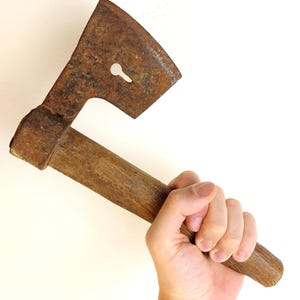 May include: A vintage hand axe featuring a weathered, rusted metal head with a distinct keyhole-shaped cutout in the center. The tool has a cylindrical wooden handle with a dark, aged patina, showing signs of extensive use and wear over time.