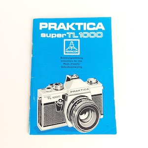 May include: A blue instruction manual for a Praktica Super TL1000 camera. The cover features the camera's image in black and white, along with the brand name and model. The manual includes instructions in multiple languages.