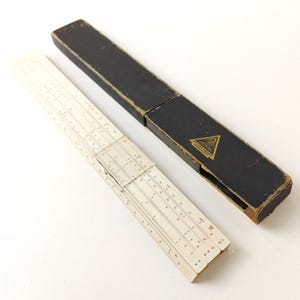 May include: A white slide rule with a black case. The slide rule has multiple scales and is labelled "K&E" on the case.